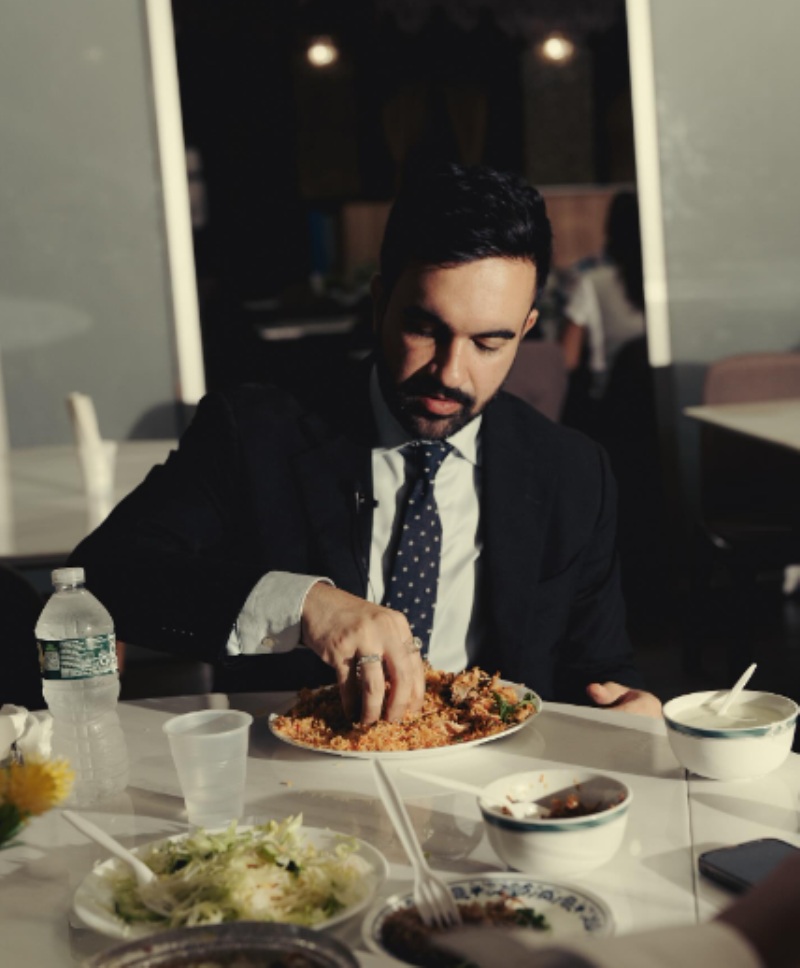 Zohran Mamdani picking up some biryani and rice with his right hand, looking down at his bowl as he does so