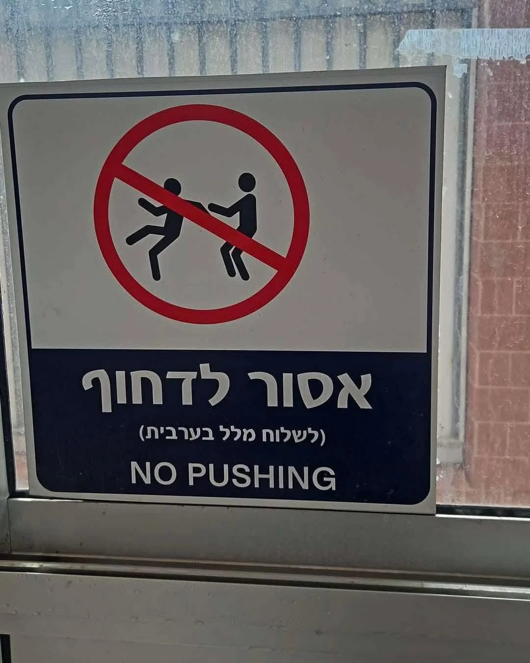 A warning sign with one figure pushing another, a red warning circle, some Hebrew writing, and 'NO Pushing' in English